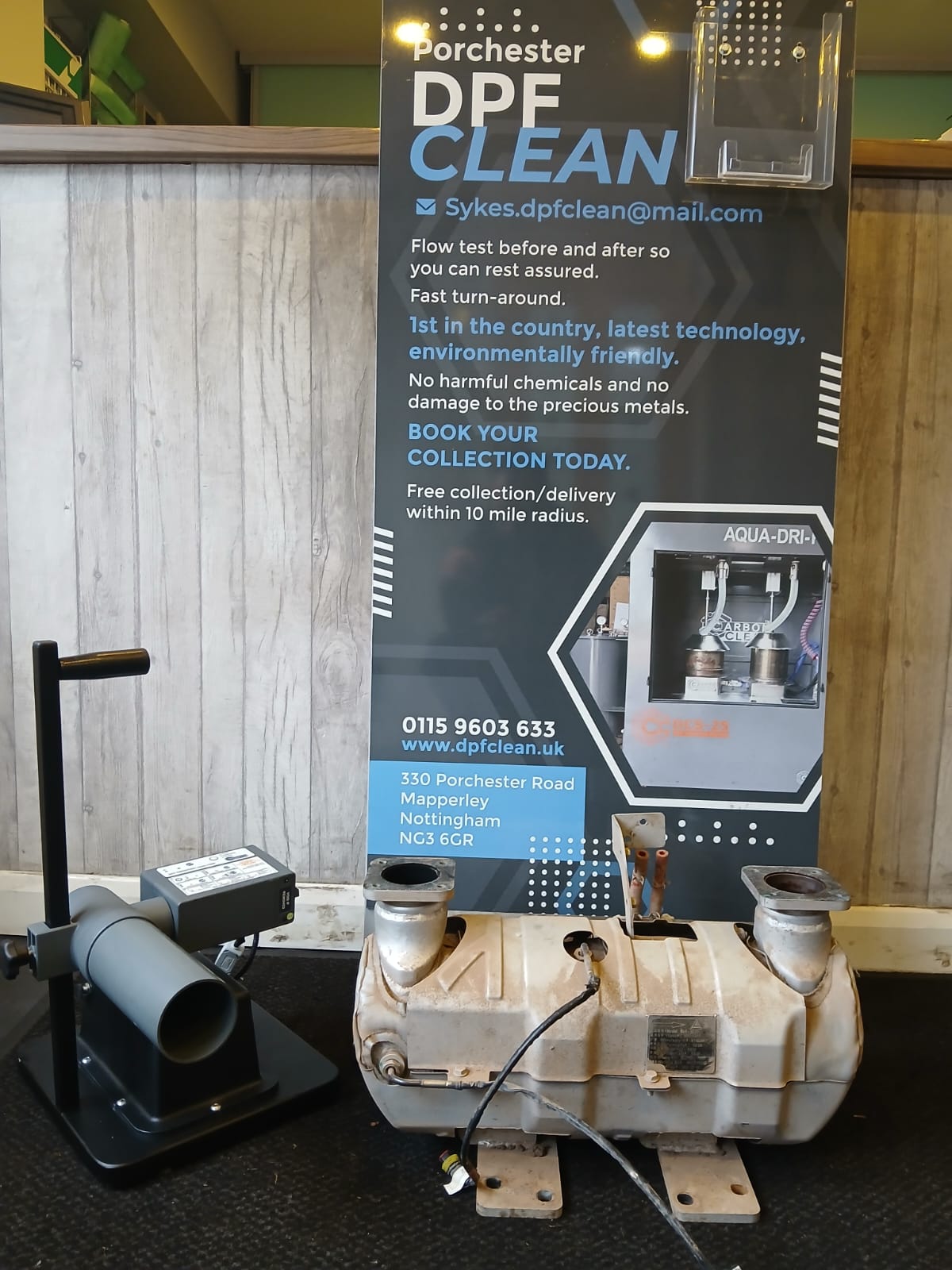 A DPF cleaning service advertisement is displayed behind a large exhaust device and a testing machine on a black surface.